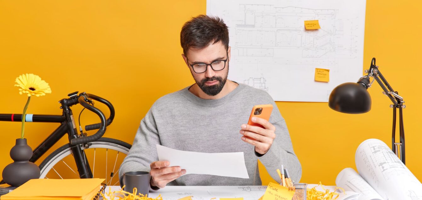 serious-bearded-european-man-concentrated-paper-uses-mobile-phone-makes-drawings-construction