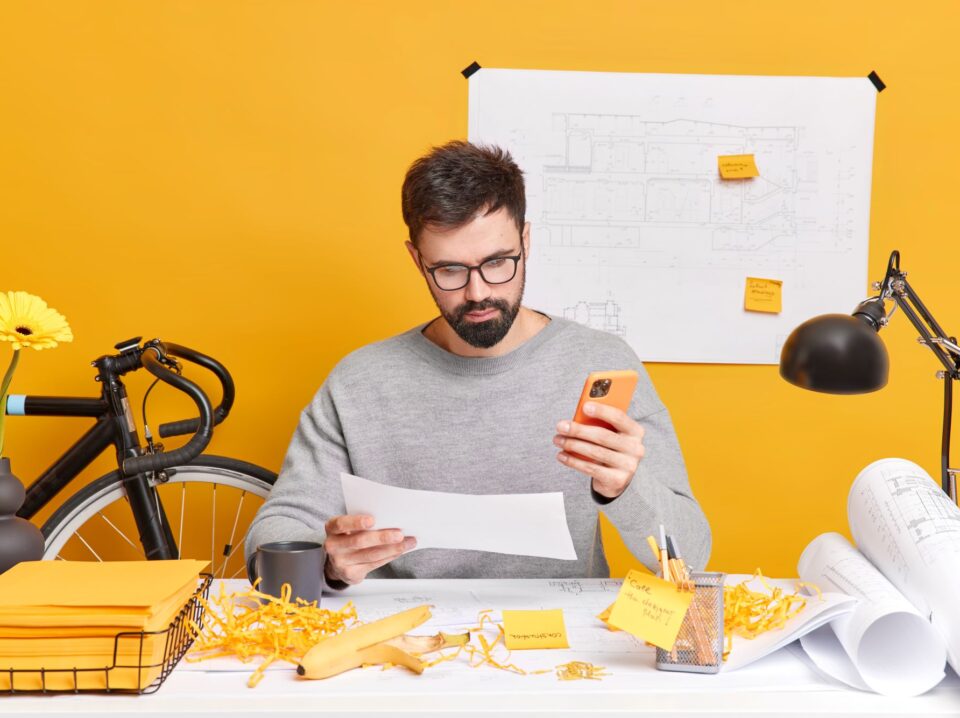 serious-bearded-european-man-concentrated-paper-uses-mobile-phone-makes-drawings-construction